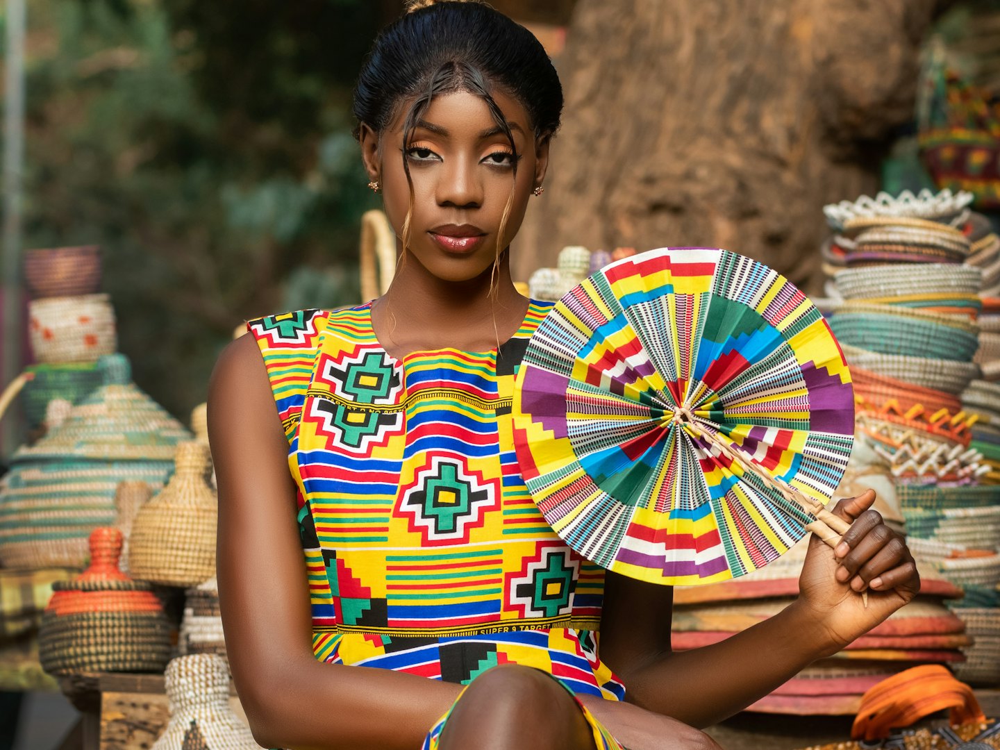 A woman in a kente-style dress holding a woven fan, with straw baskets stacked behind her.