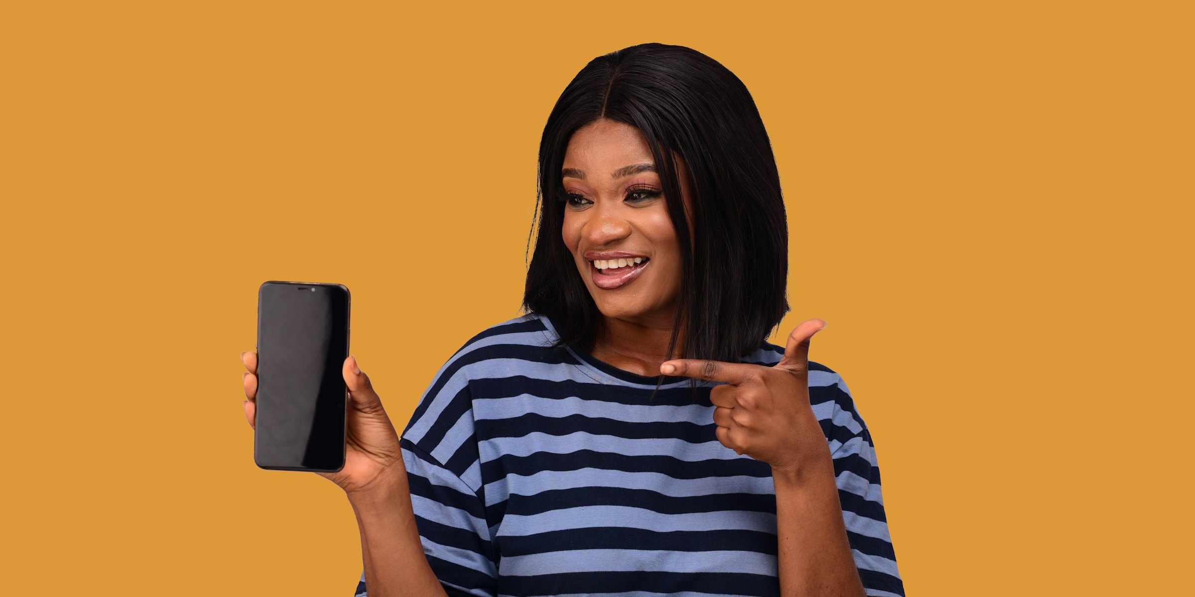 A smiling woman holding up a phone and pointing at it, against a mustard-yellow wall.