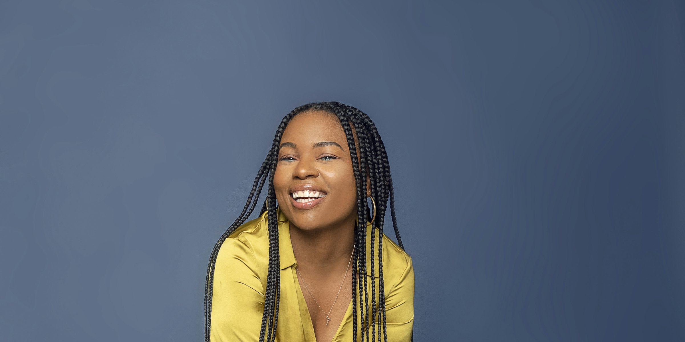 A smiling woman with long braids in a yellow satin blouse, against a blue-grey wall.
