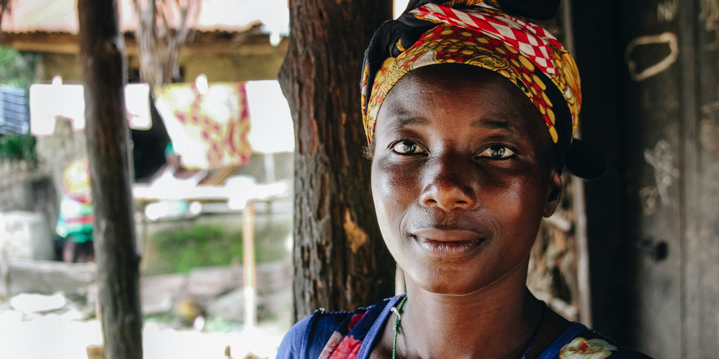 A smiling woman in a red and gold print headwrap and a floral top, standing by a tree.