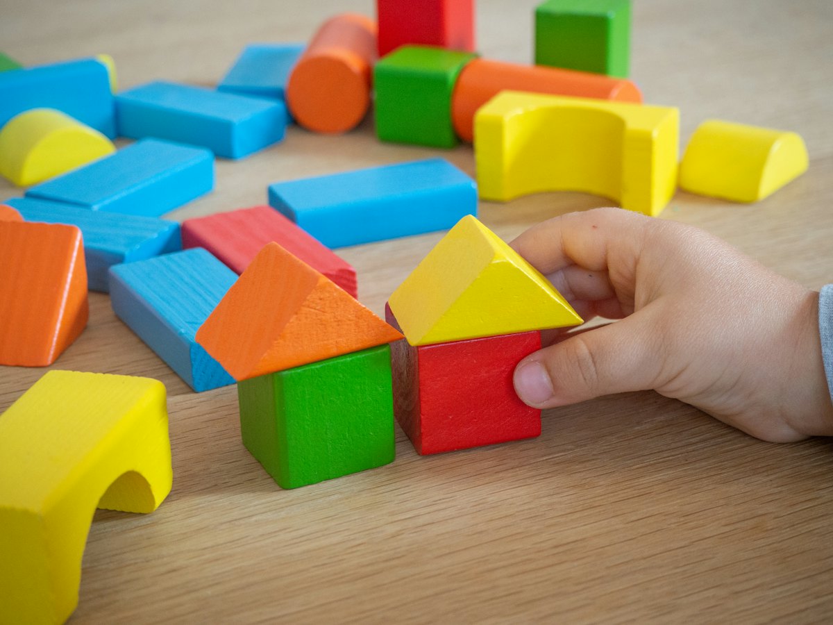 A child's hand building a house from colourful wooden blocks.